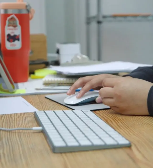 Hands using a computer mouse and keyboard at a busy desk.