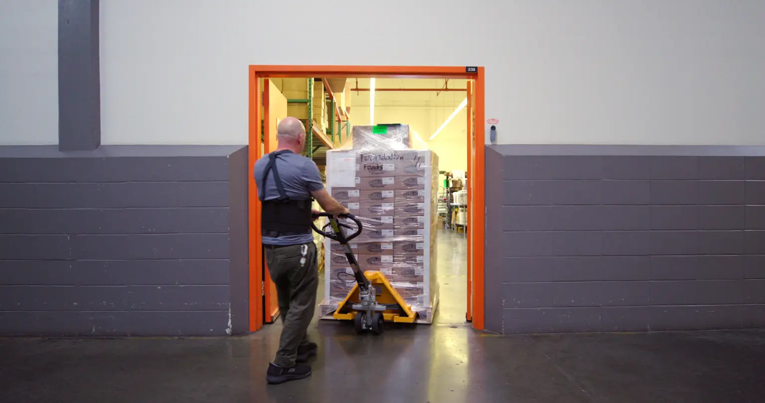 Worker operating hand truck to move stacked pallets of organic food products through warehouse door.