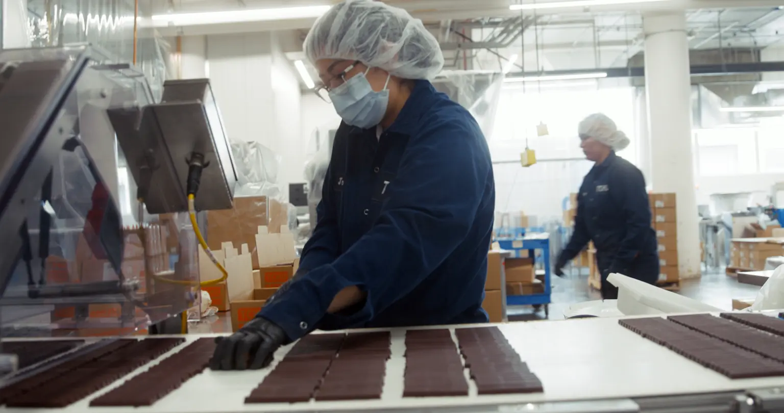 Food processing worker in hair net and protective jacket working at chocolate manufacturing vat.