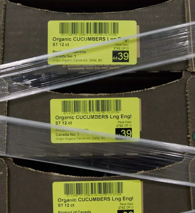 Stacked shipping cases of organic cucumbers under clear strapping, each carrying a yellow lot label with barcode, pack date and origin.