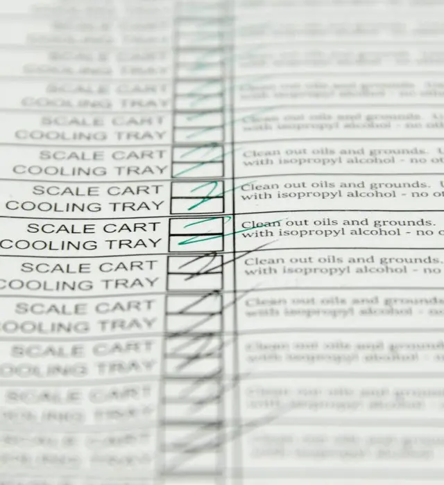 Close-up of a paper cleaning log, its rows reading SCALE CART and COOLING TRAY against the instruction to clean out oils and grounds with isopropyl alcohol, each row's checkbox marked off in pen.