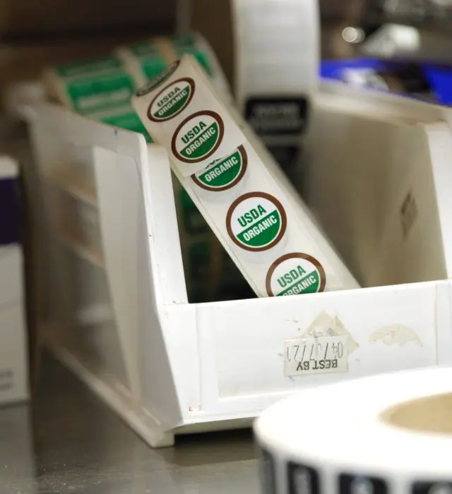 Roll of USDA Organic sticker labels in a white parts bin on a packing bench.