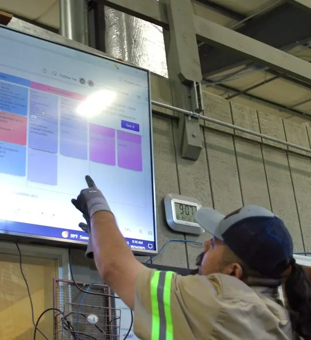 A worker in a hi-vis jacket and ball cap reaches up to point at a wall-mounted screen showing a colour-coded digital production planning board, a temperature display on the wall beside it.
