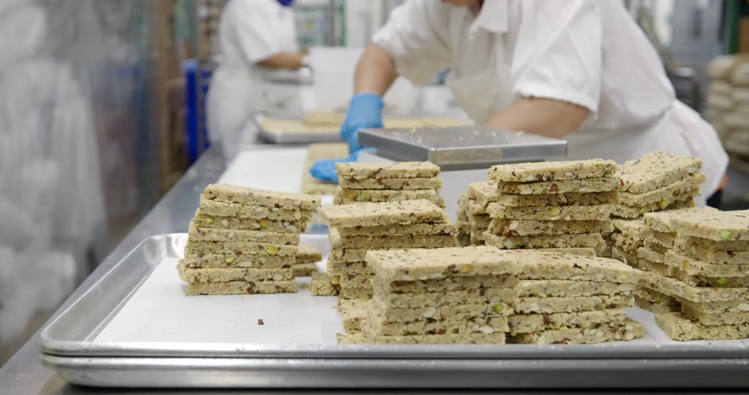 Workers in white uniforms and blue gloves handling stacks of granola bars on trays