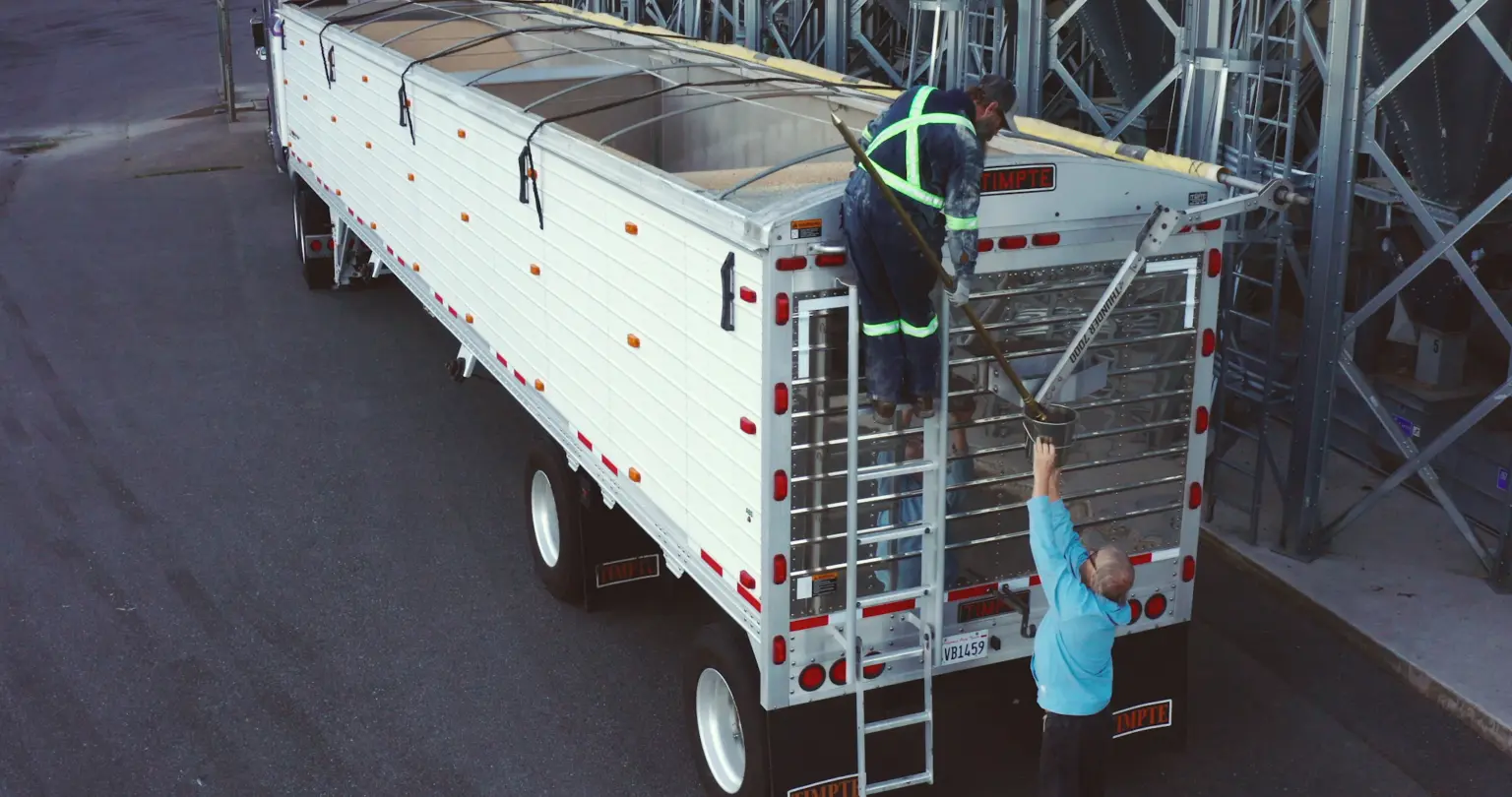 Two workers drawing a grain sample with a long probe from the open top of a hopper trailer parked beside silo loading equipment