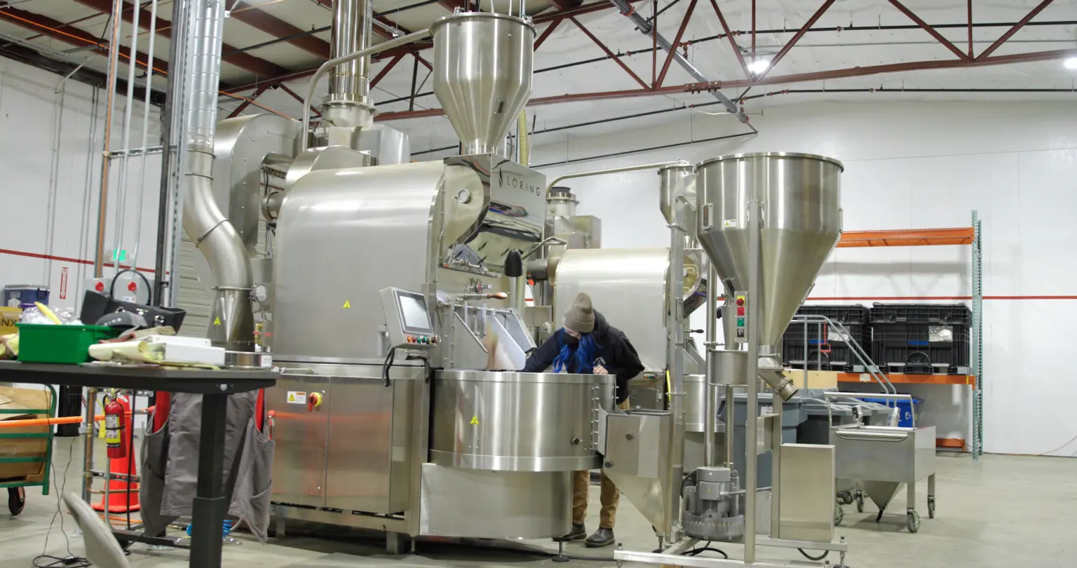 Worker operating a stainless steel coffee roaster and hopper system on a processing floor