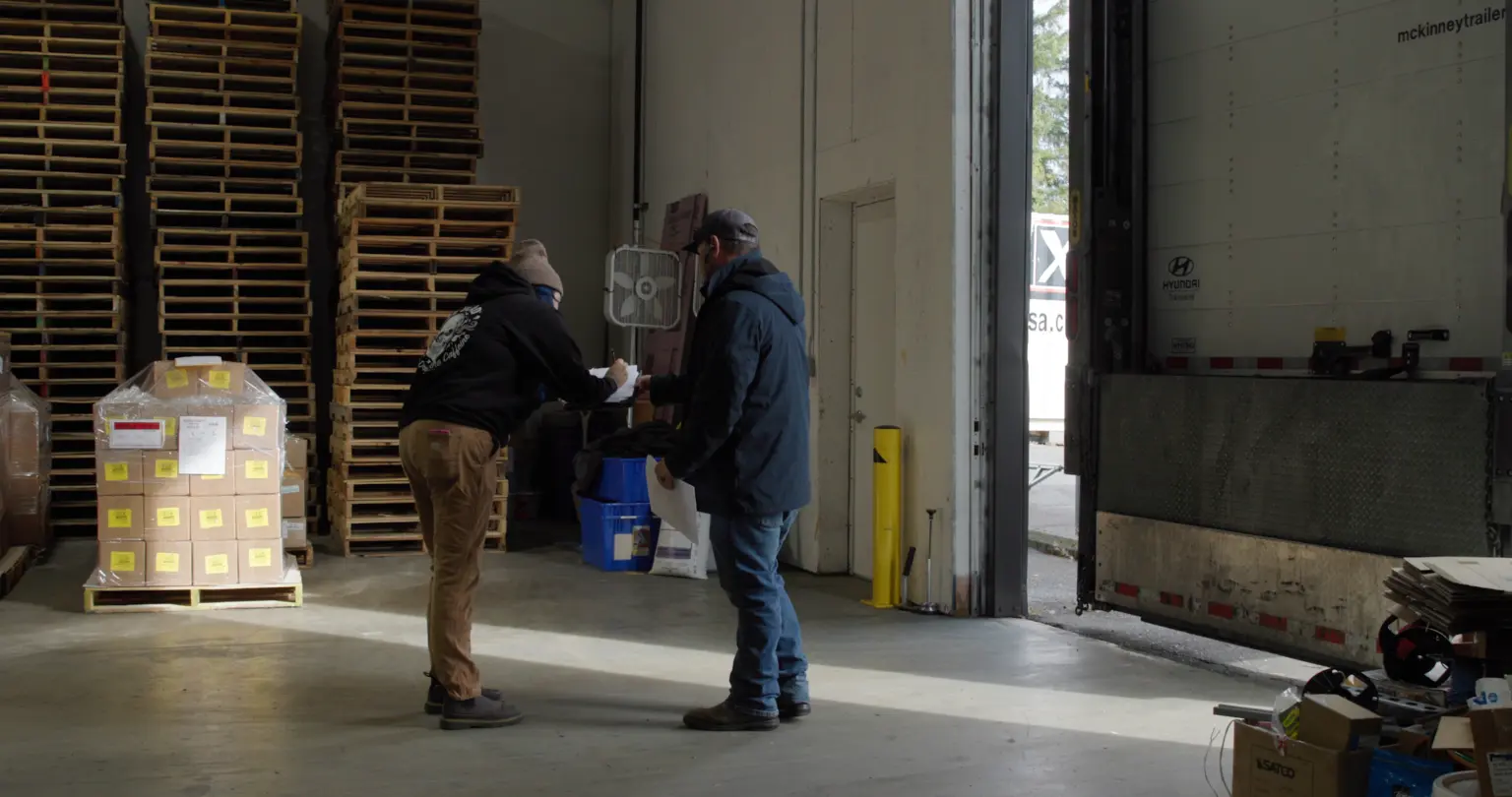 Two workers checking paperwork at a receiving dock beside a labeled pallet of cartons, with a trailer backed into the open door