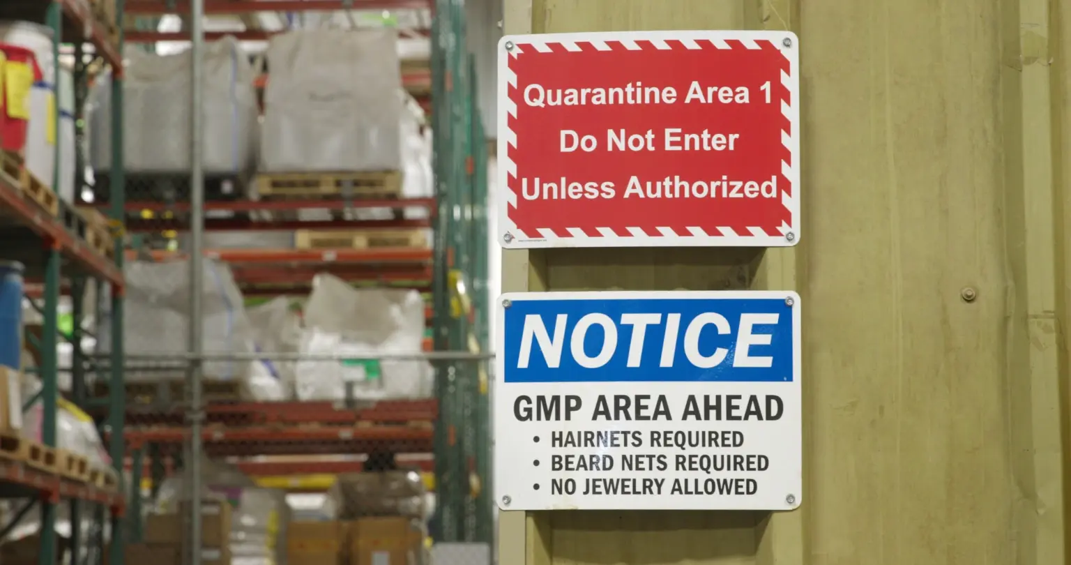 Red Quarantine Area 1 sign above a blue GMP Area Ahead notice, mounted on a post beside warehouse racking