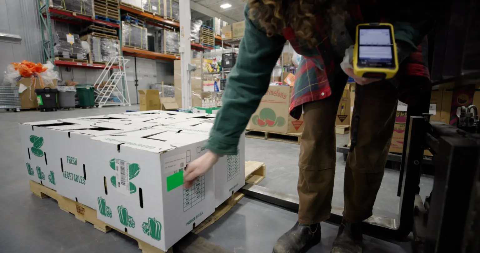 Warehouse worker scanning a produce carton marked with a green sticker beside a pallet jack