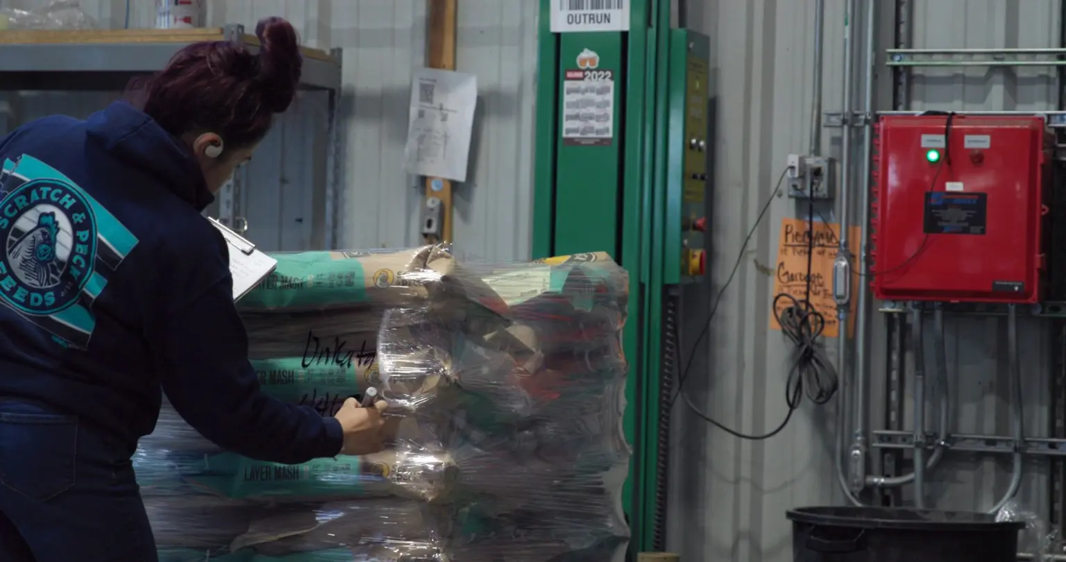Worker in a feed company sweatshirt holding a clipboard and hand-marking a shrink-wrapped pallet of layer mash sacks