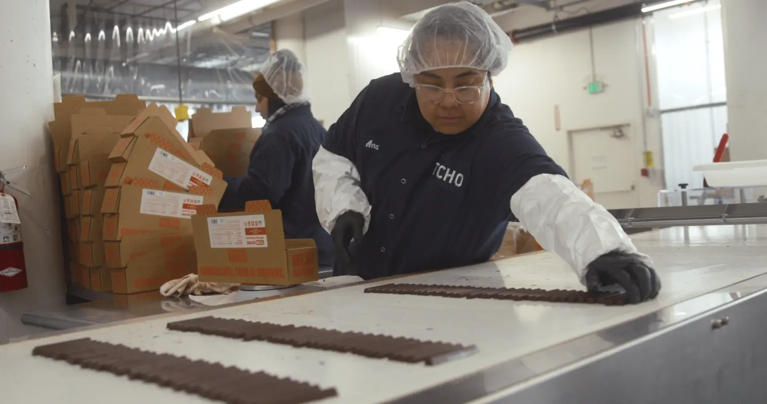 Worker in protective clothing arranging rows of chocolate batons on a food-production table