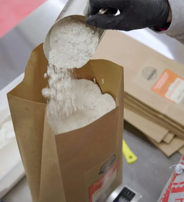 A gloved worker pouring a powdered ingredient into a bag on a scale.